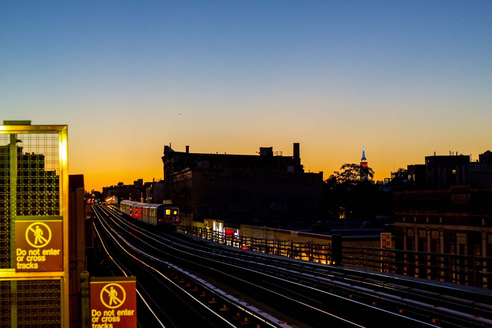 An elevated subway train arriving at a station at sunset, with the Empire State Building in the distance
