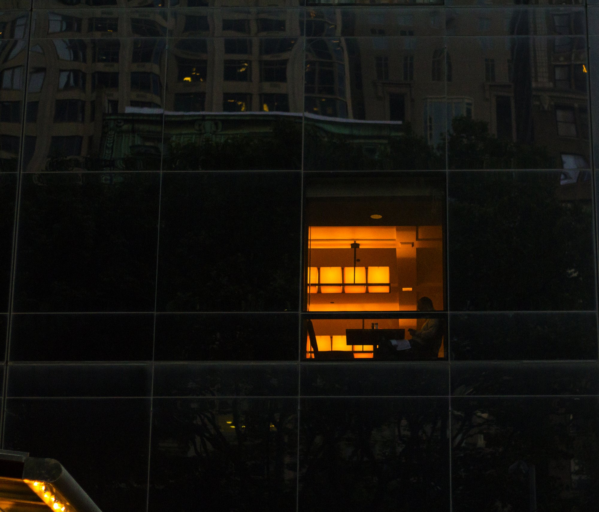 A lit office window reflected in a Manhattan skyscraper's glass facade at dusk